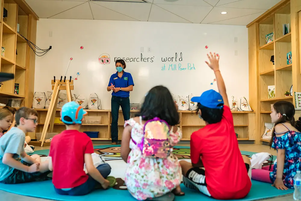 Woman wearing mask addresses children seated on classroom floor