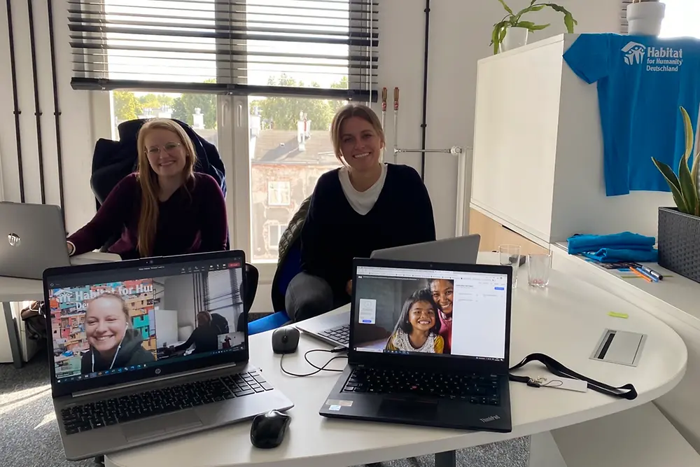 Two women are sitting at the desk in a meeting, two other women are connected virtually via laptop.