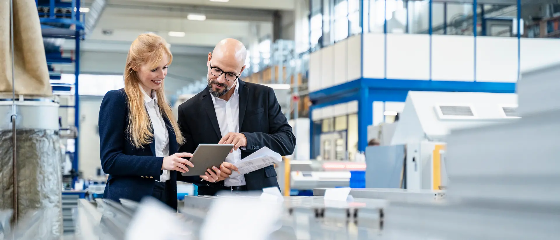 Two business people stand in a manufacturing site and look at a document