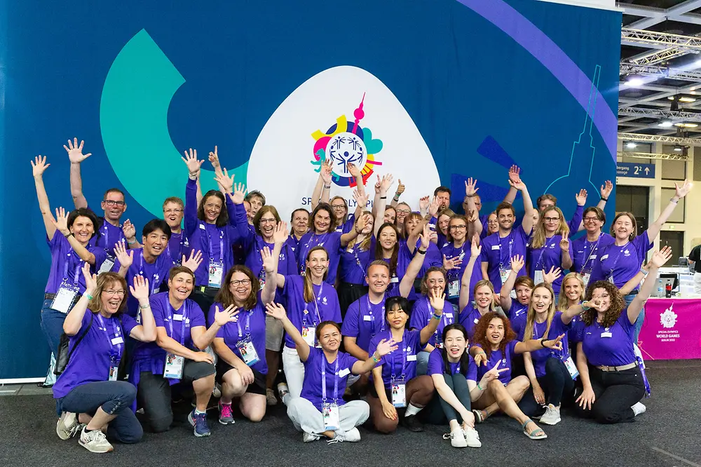 The international team of 60 Henkel volunteers wear their volunteer t-shirts and stand cheering in front of the Special Olympics World Games banner in Berlin.