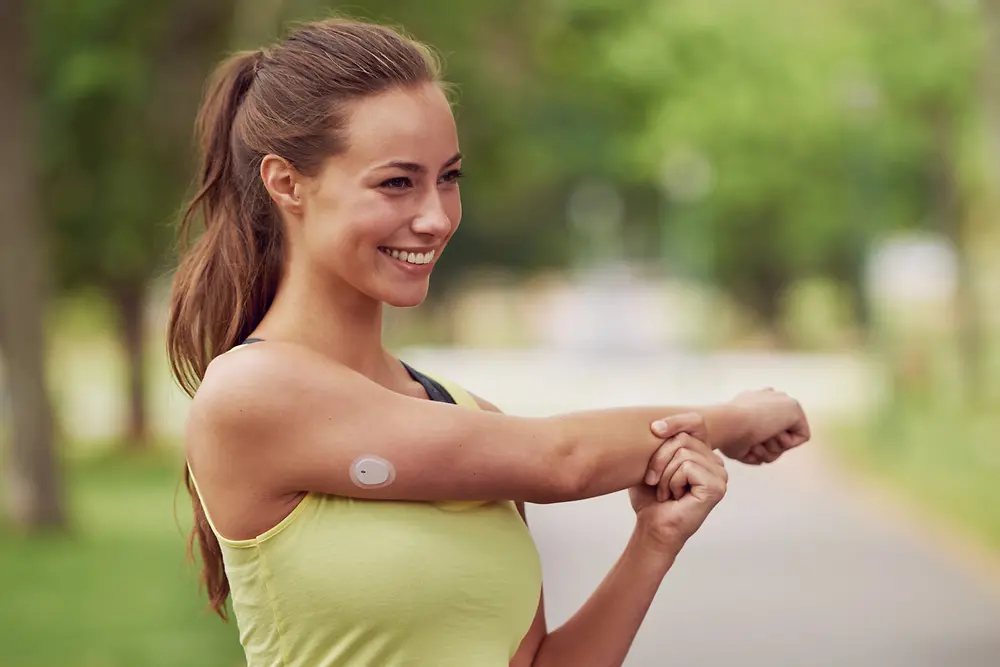 A young woman is stretching her arm preparing for exercise with a continuous glucose monitoring patch attached to her upper right arm.