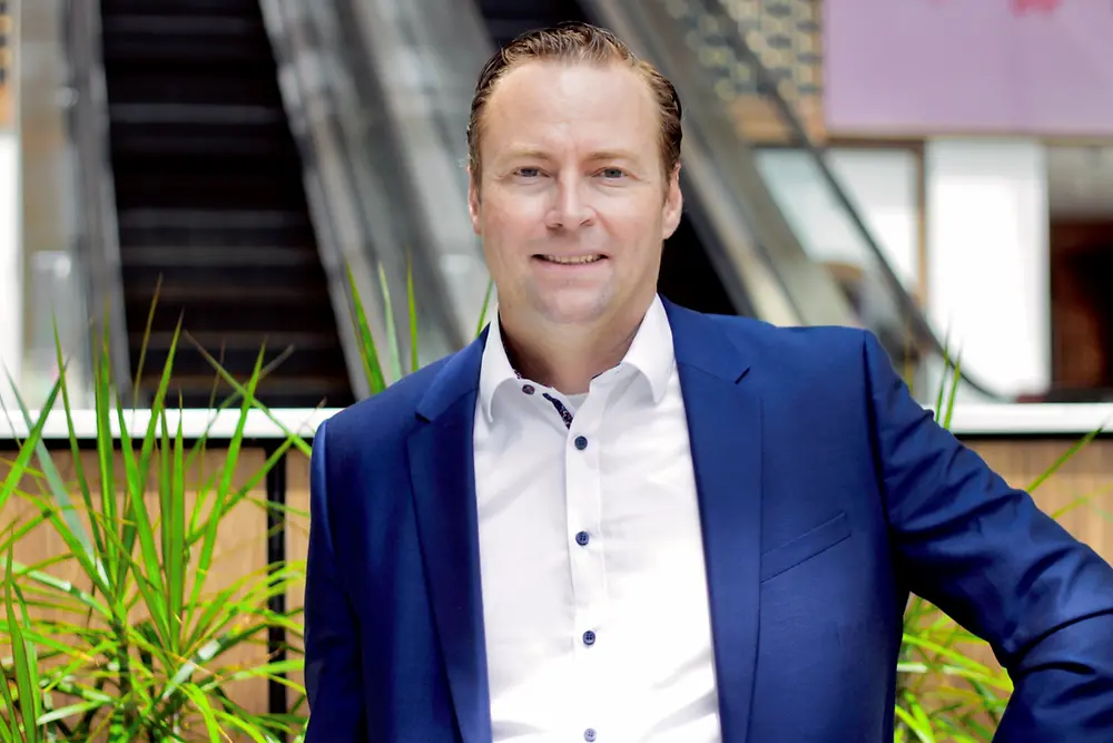 Man in a blue suit smiles in front of greenery in an office building