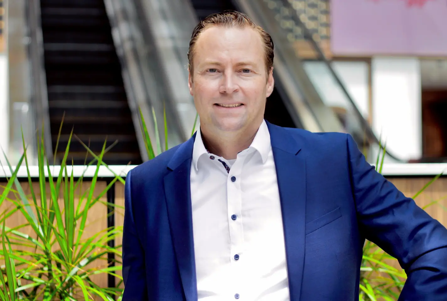 Man in a blue suit smiles in front of greenery in an office building