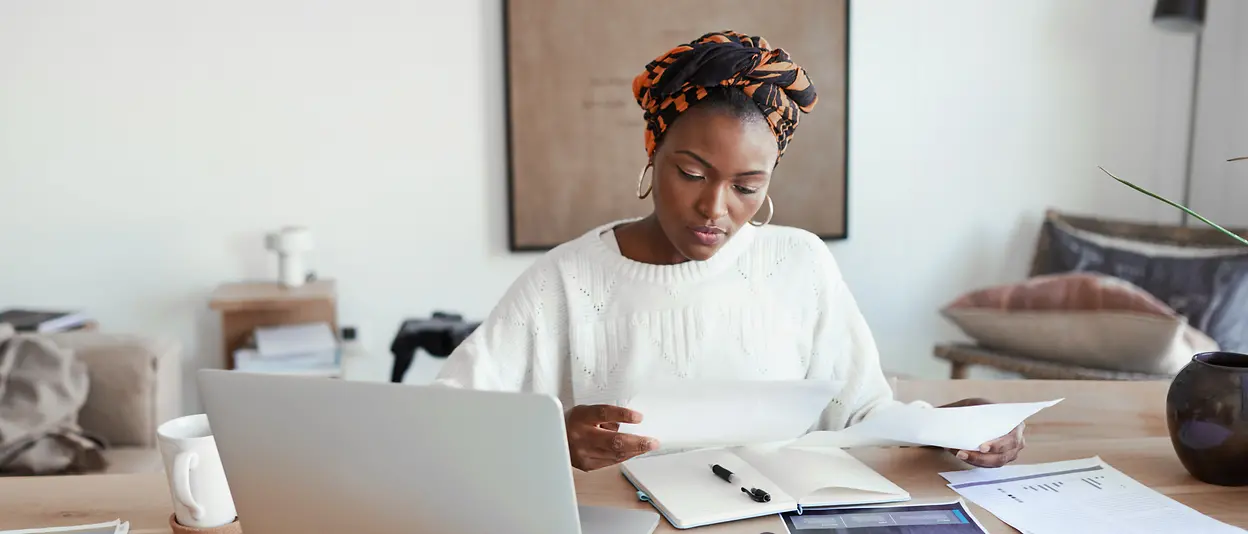 A woman sits at her desk at home and looks at documents.