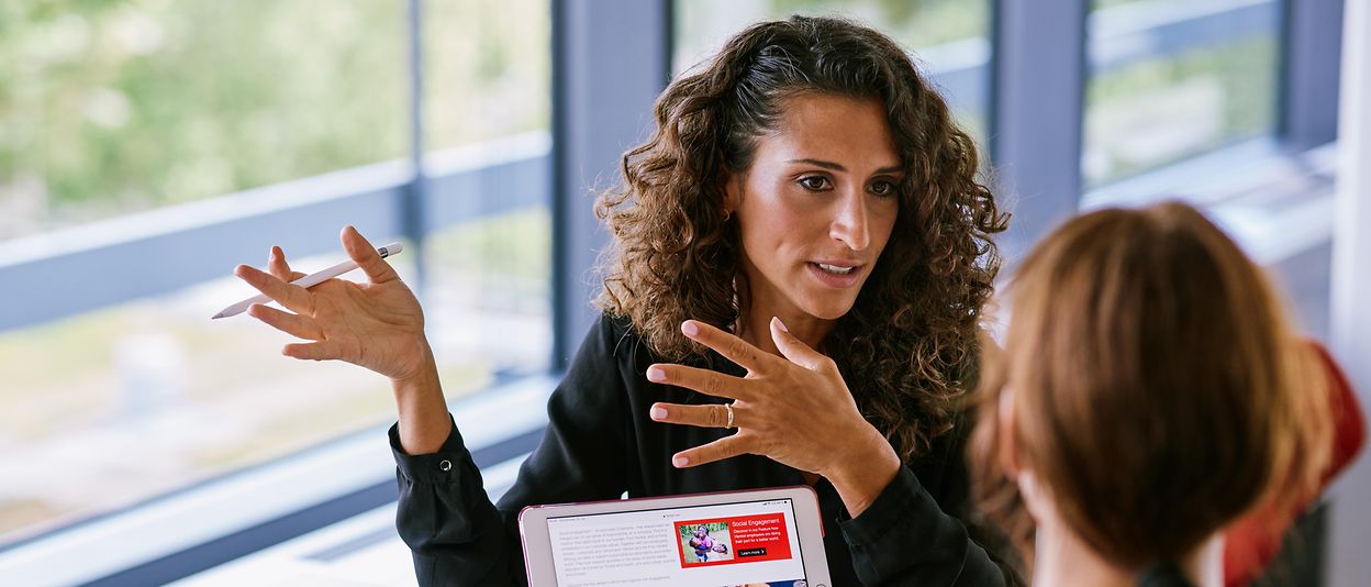 A woman explains something to another woman and shows her a presentation on her tablet