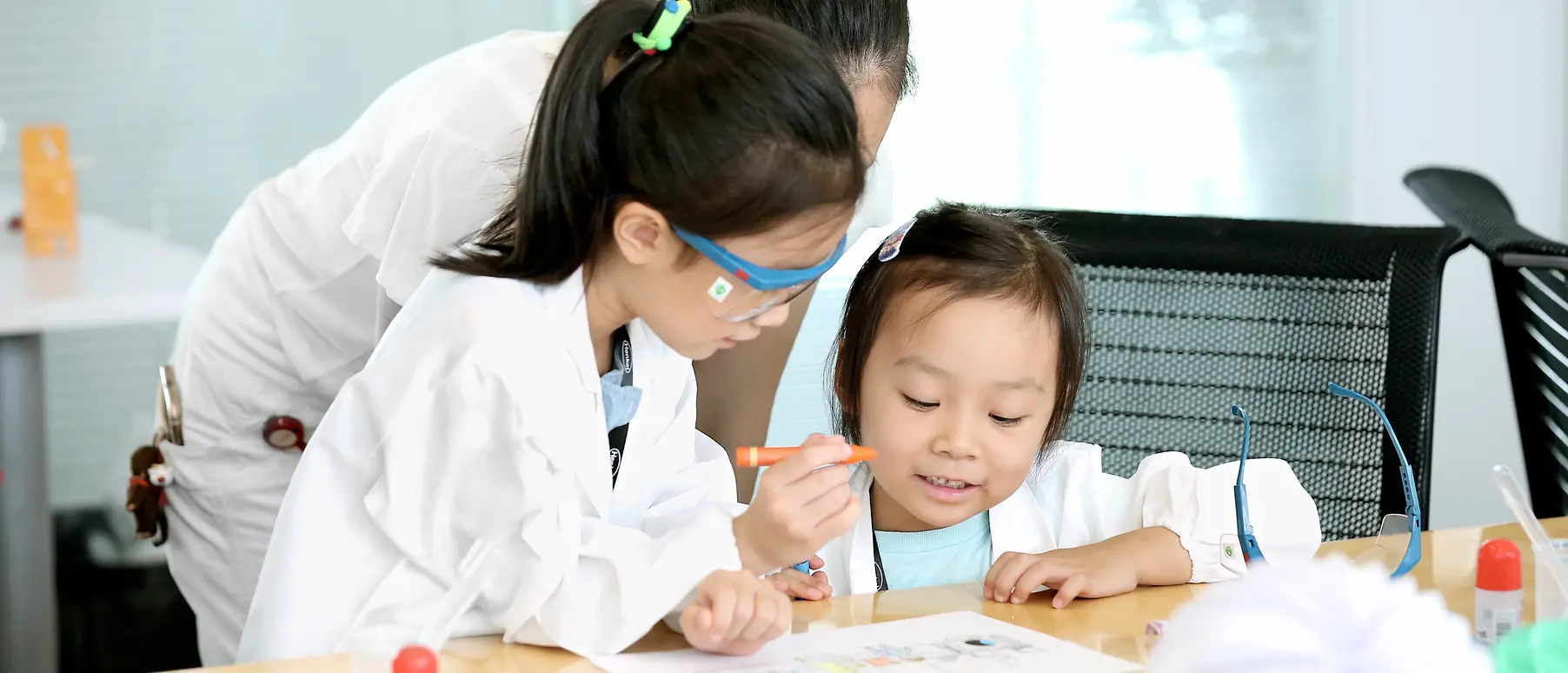 Two children and a woman in a research coat colour a picture at a table