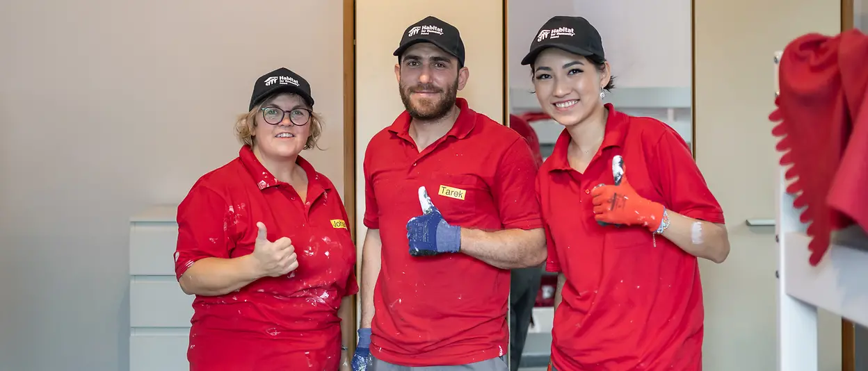 Three volunteers in construction clothing and matching t-shirts give a thumbs up and smile into the camera.