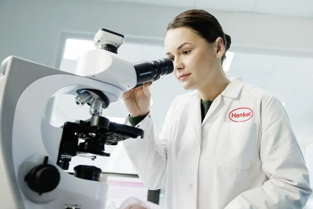 Eine Frau im Laborkittel mit Henkel-Logo schaut durch ein Mikroskop